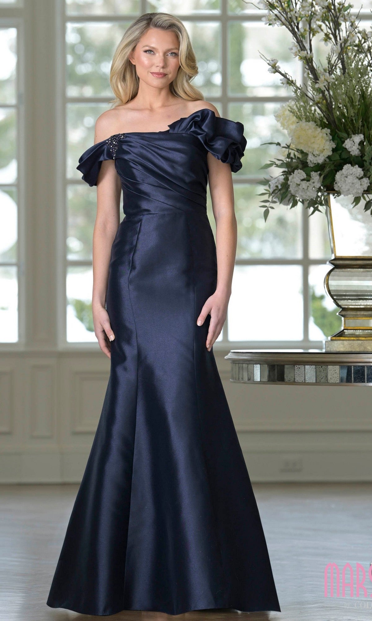 A woman stands indoors near a table with a large floral arrangement, wearing the Long Formal Dress: Marsoni MV1407 in midnight—a ruffled off-shoulder, floor-length evening gown with a mermaid silhouette.